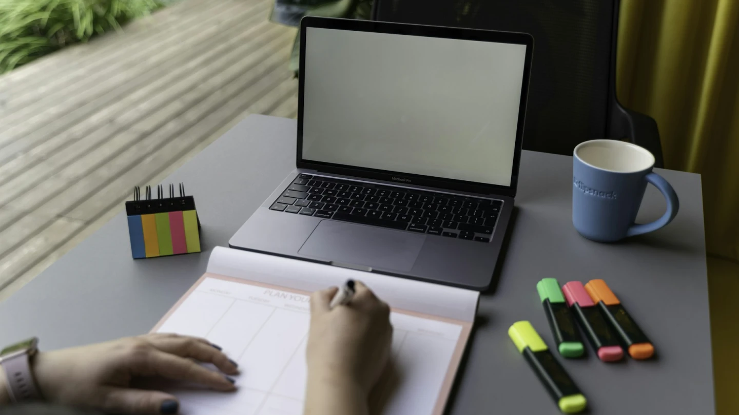 A person reviewing a planner beside a laptop and organized desk supplies.