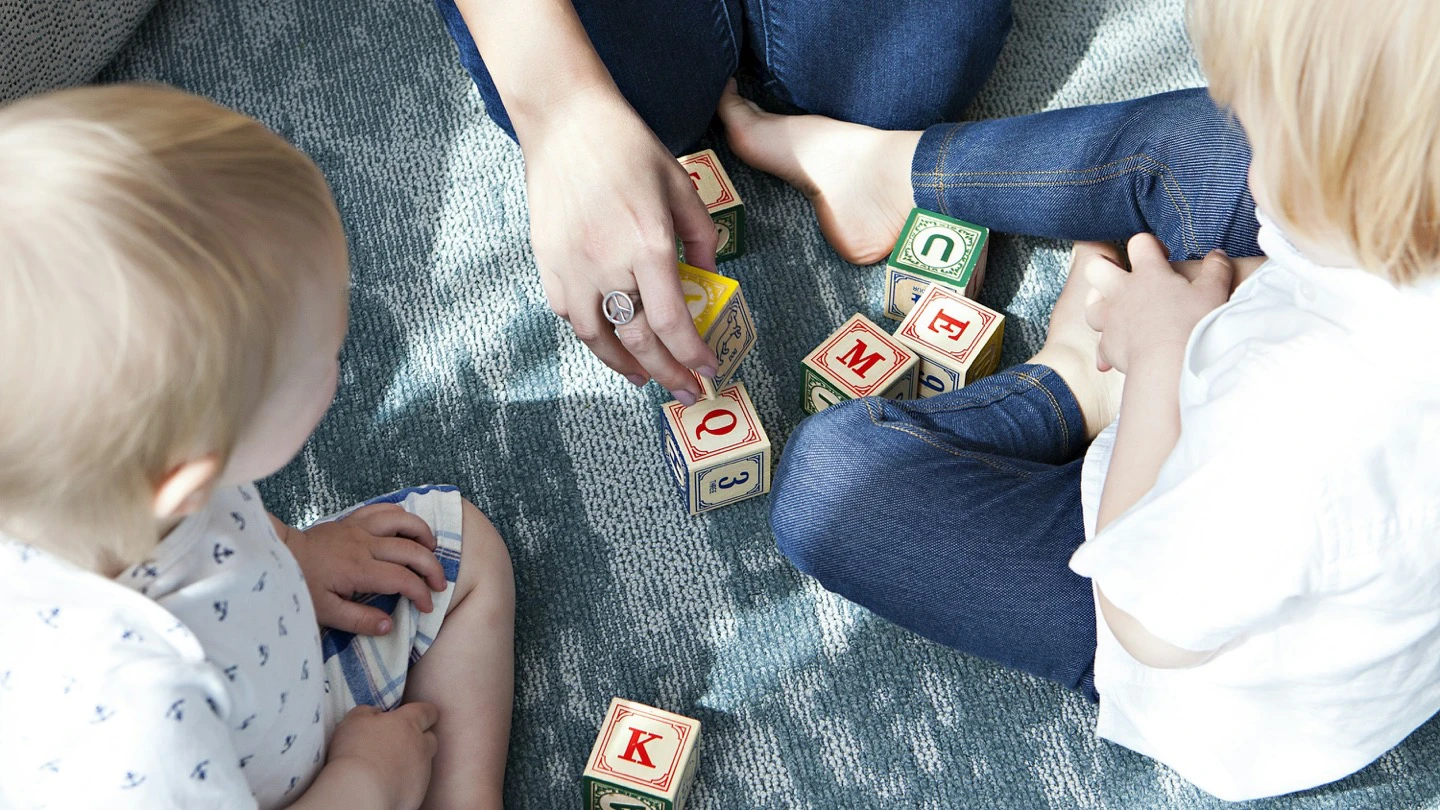 Two young children playing with wooden letter blocks on a blue rug.